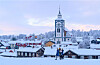 Snötäckt stad med kyrka, trähus och två personer som går i förgrunden.