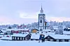Snötäckt stad med kyrka, trähus och två personer som går i förgrunden.