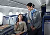 Flygvärdinna i grå uniform serverar en kvinna som sitter i ett flygplansäte och håller en kopp.