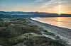 Soluppgång över en strand med lugnt vatten, sanddyner och berg i bakgrunden.