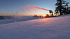 Snökanoner sprutar snö i en skidbacke i skymningen med träd och orange himmel i bakgrunden.