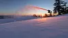 Snökanoner sprutar snö i en skidbacke i skymningen med träd och orange himmel i bakgrunden.