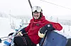 En person i röd skidjacka sitter i en lift med snöiga backar och dimma bakom.