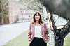 En kvinna står utomhus bredvid ett träd med byggnader och en väg i mjuk fokus bakom.