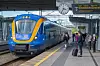 Blått och gult regionaltåg står vid en perrong med resenärer och en avgångstavla i bakgrunden.