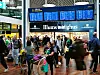 Människor går genom en stationshall med stora blå avgångstavlor och butiken Illums Bolighus i bakgrunden.