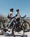 Två personer med cyklar står vid en stig nära havet under en klar blå himmel.