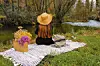 Person med stråhatt sitter på filt vid en flod med picknickkorg och blommor i gräset.