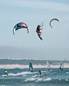 Flera personer utövar kitesurfing och windsurfing i vågigt vatten nära en strand.