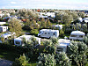 Husvagnar och campingvagnar står bland träd på den gröna campingplatsen Insel Camping Borkum, som förvärvats av First Camp, med hus i bakgrunden.