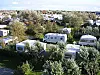 Husvagnar och campingvagnar står bland träd på den gröna campingplatsen Insel Camping Borkum, som förvärvats av First Camp, med hus i bakgrunden.