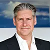 Johan Lundgren i svart kavaj och vit skjorta står framför en blå och vit bakgrund.