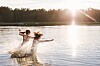 Två personer hoppar i vattnet i en sjö i Finland med midnattssolen och skog i bakgrunden.