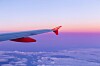 Flygplansvinge med röd vingspets ovanför moln vid solnedgång.
