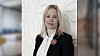 En kvinna i svart blazer och vit blus står framför en ljus, dekorativ vägg.