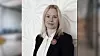 En kvinna i svart blazer och vit blus står framför en ljus, dekorativ vägg.