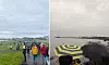 En folksamling står på en gräsmatta under molnig himmel; flera personer håller paraplyer.