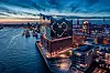 Flygbild över Hamburgs upplysta hamn, vatten och täta stadsbebyggelse i skymningen.