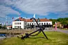 Vit byggnad med röda tak vid en kaj, flaggor och ett stort ankare på gräset.