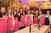 A group of smiling hospitality professionals in pink aprons pose behind a dining table at an indoor event.