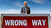 En man står bakom en röd skylt med texten ”WRONG WAY” mot en blå himmel.