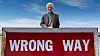En man står bakom en röd skylt med texten ”WRONG WAY” mot en blå himmel.
