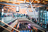 Resenärer i en modern flygterminal med rulltrappor, glasräcken och informationsskärmar.