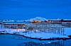 Snötäckt kustby med fiskställningar, vatten och fjäll i bakgrunden.