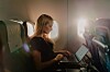 En kvinna arbetar vid en laptop i ett flygplans kabin, med solljus från två fönster.