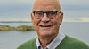 Porträtt av en äldre man med glasögon framför ett lugnt vattenlandskap.