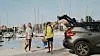 Två resenärer står bredvid en grå bil vid en marina med segelbåtar.