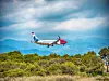 Ett passagerarflyg flyger över grön vegetation med blå berg och moln i bakgrunden.