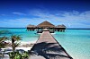 Träbrygga över turkost vatten leder mot bungalowbyggnader på en solig maldivisk resort.
