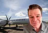 Leende man framför ett turbopropflygplan på en flygplats under en molnig himmel.