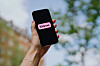 En hand håller upp en svart mobiltelefon med Klarnas rosa logotyp mot en suddig utomhusbakgrund.