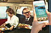 En smartphone med texten ”Find your wave” hålls framför två resenärer som äter i en äldre flygplanskabin.