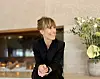 Leende kvinna i svart klädsel sitter i en hotellmiljö med en blomsterbukett bakom sig.