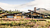 Låg hotellbyggnad omgiven av blommande vegetation, barrskog och fjäll under en ljus himmel.