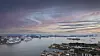Panoramavy över en hamn med fartyg, småbåtar och stadsbebyggelse under en färgstark kvällshimmel.