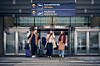 Tre resenärer med bagage står framför en flygplatsterminal med skyltar för ankommande och avgående.