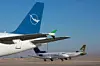 Två passagerarflygplan på en flygplats; det främre har blå stjärtfena med vit fågellogotyp.