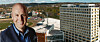 Leende man i blå skjorta framför ett stort hotell och ett stadslandskap.